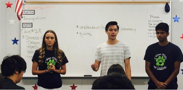 A girl and two boy high school students stand at the front of a classroom, talking about distracted driving. A girl and two boy high school students stand at the front of a classroom, talking about distracted driving.